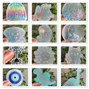 May include: A set of 10 iridescent holographic stickers featuring various designs, including a rainbow, a ghost, a moon, a sun, a spider, a dinosaur, an evil eye, a cat, a dog, and an astronaut.  The stickers are printed on clear vinyl and have a holographic finish.
