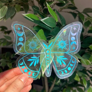 May include: A clear holographic sticker of a butterfly with celestial designs. The butterfly has a blue iridescent finish and features moons, stars, and suns.