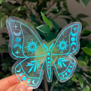 May include: A holographic sticker of a butterfly with celestial designs. The butterfly is outlined in blue and has a pattern of moons, stars, and suns on its wings.