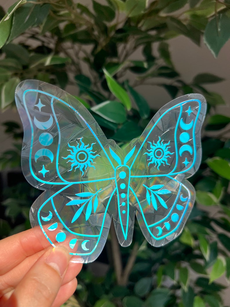 May include: A holographic sticker of a butterfly with celestial designs. The butterfly is outlined in blue and has a pattern of moons, stars, and suns on its wings.