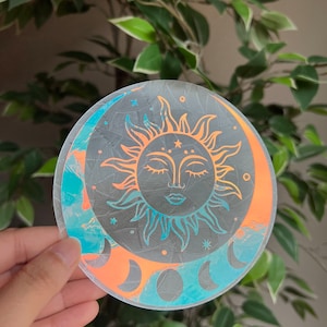 May include: A holographic sticker featuring a sun and moon design. The sun is orange and yellow, and the moon is blue and green. The sticker is round and has a cracked glass effect.