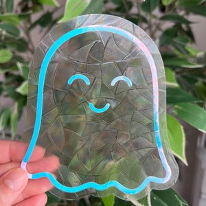 May include: A clear, ghost-shaped sticker with a smiling face and a blue outline. The sticker has a textured, iridescent surface and is held in front of a background of green leaves.