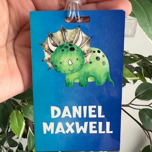 Peut inclure: Une étiquette de bagage bleue avec une illustration de triceratops vert. L'étiquette porte le nom "DANIEL MAXWELL" en lettres blanches. Une boucle en plastique transparent est fixée en haut pour l'attacher aux bagages ou au sac à dos.