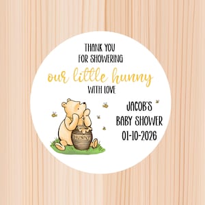 May include: A round white sticker featuring a cartoon bear sitting on green grass next to a honey pot. The text reads: THANK YOU FOR SHOWERING our little hunny WITH LOVE, JACOB'S BABY SHOWER, and 01-10-2026. Small bees are flying around the bear.