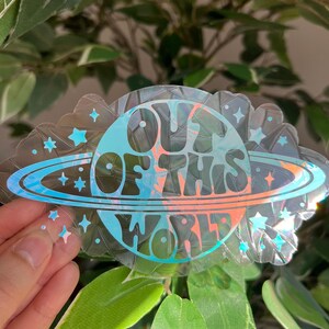 May include: A clear sticker with the words "OUT OF THIS WORLD" in a retro font. The design features a planet with rings and stars in shades of blue, orange, and pink. The sticker has a scalloped edge and holographic accents.