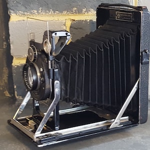 May include: A black vintage camera with a bellows system and a silver metal frame. The camera has a lens on the front and a handle on the top.
