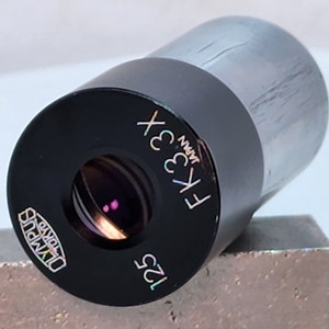 May include: A black microscope objective lens with the text "FK 33x" and "125" printed on it. The lens is made by Olympus and is from Japan.