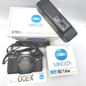 Puede incluir: Una cámara Minolta X-700 negra de 35 mm con tapa de objetivo y correa para el cuello, junto con un flash y dos cajas. Una caja dice "MINOLTA X-700" y la otra "MINOLTA 50/1.4 MD".