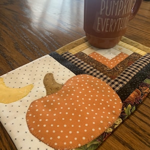 May include: A burnt orange ceramic mug with the white text PUMPKIN EVERYTHING sits on a rectangular quilted fabric mat featuring a white background with black crosses, a yellow crescent moon, and an orange polka-dot pumpkin applique.