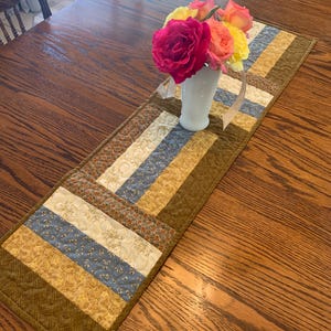 May include: A rectangular quilted table runner featuring a patchwork design of alternating strips in shades of olive green, dusty blue, cream, and floral patterns. A white vase containing a bouquet of pink, yellow, and orange roses sits on top.