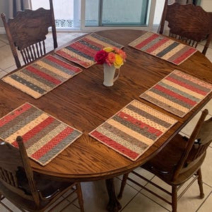 May include: An oval wooden dining table with a dark brown finish, surrounded by four matching wooden spindle-back chairs. Five rectangular fabric placemats with red, grey, and cream striped patterns are arranged on the table surface.