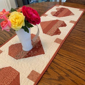 May include: A white milk glass pitcher filled with yellow and pink roses sits on a rectangular quilted table runner. The runner features a cream-colored background with a repeating pattern of red heart shapes made from various fabric strips and textures.