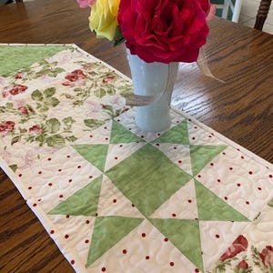 May include: A quilted table runner featuring a patchwork design with sections of floral rose patterns and a central green star motif on a white background with small red dots. A white vase holds a bouquet of vibrant red, yellow, and pink roses.