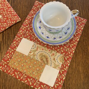 May include: A quilted fabric placemat featuring a red and green floral pattern with a central patchwork section of cream, paisley, and floral fabric. A white porcelain teacup with a gold rim and a matching saucer with blue floral accents sit on top.