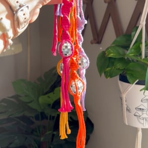 May include: A set of five colorful macrame keychains with disco ball charms. Each keychain is made with a different color of yarn: orange, pink, purple, and lavender. The charms are silver with mirrored surfaces.