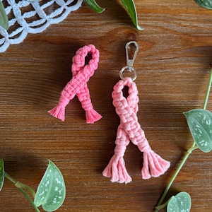May include: Two pink macrame ribbon-shaped items on a wooden surface. One is a standalone ribbon, and the other is a keychain with a silver clasp. The macrame ribbons have a textured appearance with fringed ends. Green leafy vines surround the ribbons.