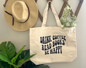 May include: A white canvas tote bag with black text that reads "Drink Coffee Read Books Be Happy" in a retro font. The bag is hanging on a wooden wall rack with a brown hat and a sprig of greenery.