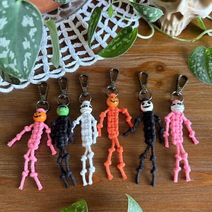 Puede incluir: Seis llaveros de esqueleto de macramé hechos a mano en rosa, negro, blanco y naranja. Cada llavero presenta una cabeza de diferente color, incluyendo una calabaza, y un cierre plateado. Los llaveros están dispuestos sobre una superficie de madera.