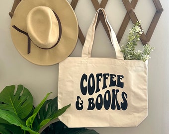 May include: A white canvas tote bag with black text that reads "COFFEE & BOOKS" in a retro font. The bag is hanging on a wooden wall rack with a brown hat and green plant visible in the background.