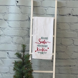 May include: White kitchen towel with red embroidered text that reads "Who needs Santa when you have KeeKee". The towel is hanging on a white wooden ladder leaning against a white brick wall. A small artificial pine tree is in the foreground.