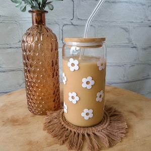 May include: A glass jar with a bamboo lid and a clear glass straw. The jar is filled with a light brown liquid and has white flower decals. The jar is sitting on a brown woven coaster with fringe.