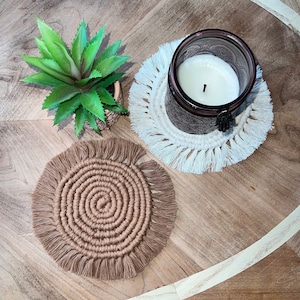 May include: Two round macrame coasters with fringe. One coaster is brown and the other is white. A candle is sitting on the white coaster.