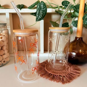 May include: Two clear glass tumblers with bamboo lids and glass straws. One tumbler has orange bow designs, the other has gold bow designs. Both are on a white surface with a brown woven coaster.