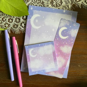 May include: Stationery set with a celestial theme, including notepads and a bookmark with a dreamy sky design, crescent moons and stars. Two pens, lavender and pink, are on a wooden surface. The set is ideal for jotting down notes or journaling.