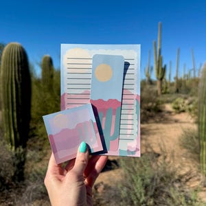 May include: A set of stationery featuring a desert landscape design. The set includes a notepad with lined pages, a bookmark, and a small square sticky note pad. The design features a blue sky, pink mountains, and cacti.