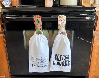 May include: Two white kitchen towels hanging on a black stove. The towel on the left has a green plant graphic and the text "just one more". The towel on the right has the text "COFFEE & BOOKS" in black.