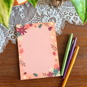 May include: A rectangular notepad with a soft pink center and a border featuring colorful autumn leaves, flowers, and acorns. Three capped pens in light green, purple, and orange are positioned to the right of the notepad on a wooden surface.