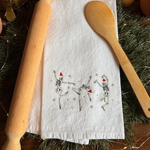 May include: A white kitchen towel with a whimsical design of dancing skeletons wearing Santa hats. A wooden rolling pin and spoon are placed on either side of the towel. The towel is surrounded by greenery and festive decorations.