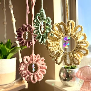 Può includere: Ornamenti floreali in macramè fatti a mano nei toni del rosa, verde e beige, appesi davanti a una finestra. Ogni fiore presenta un pendente di cristallo centrale. Decorazione bohémien.