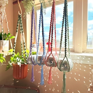 May include: A selection of colourful macrame plant hangers, each holding a disco ball planter. The hangers, in shades of green, purple, blue, and pink, are suspended from a wooden rod near a window. The display creates a decorative and eye-catching feature.