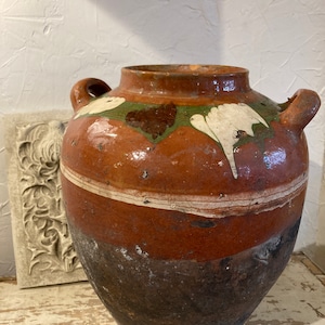 May include: A large, brown, earthenware pot with white and green painted designs. The pot has two handles and a wide opening. The pot is slightly weathered and has a dark brown glaze.