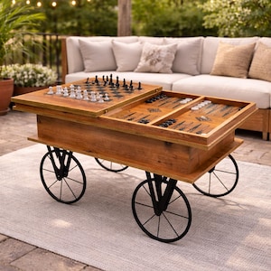 May include: A wooden game table on wheels, featuring a chess board and a backgammon board. The table has a natural wood finish and black metal wheels. The table is set on a light gray rug, with a beige sofa in the background.