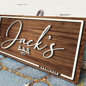 May include: A rustic wooden sign with a white border and the text "Jack's Bar Est. 2018" in white lettering.