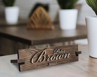 May include: A wooden desk nameplate with the text "Peter Brown - Manager" engraved in a cursive font. The nameplate is brown and has a natural wood grain finish.