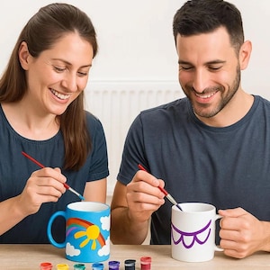 May include: Two people painting ceramic mugs. One mug is blue with a rainbow and sun design, the other is white with a purple design. Small paint pots in various colours are on the table.