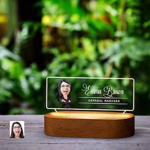 以下が含まれることがあります： 木製の台座に、透明なアクリル製のプレートが置かれ、プレートには人物の漫画風ポートレートと「Emma Brown, General Manager」というテキストが描かれています。プレートは下から照明されています。