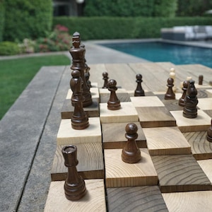 Puede incluir: Un juego de ajedrez de madera con piezas de madera marr&oacute;n oscuro y clara sobre un tablero &uacute;nico y elevado. El tablero est&aacute; instalado al aire libre cerca de una piscina y un c&eacute;sped verde. Las piezas de ajedrez est&aacute;n en las posiciones iniciales.