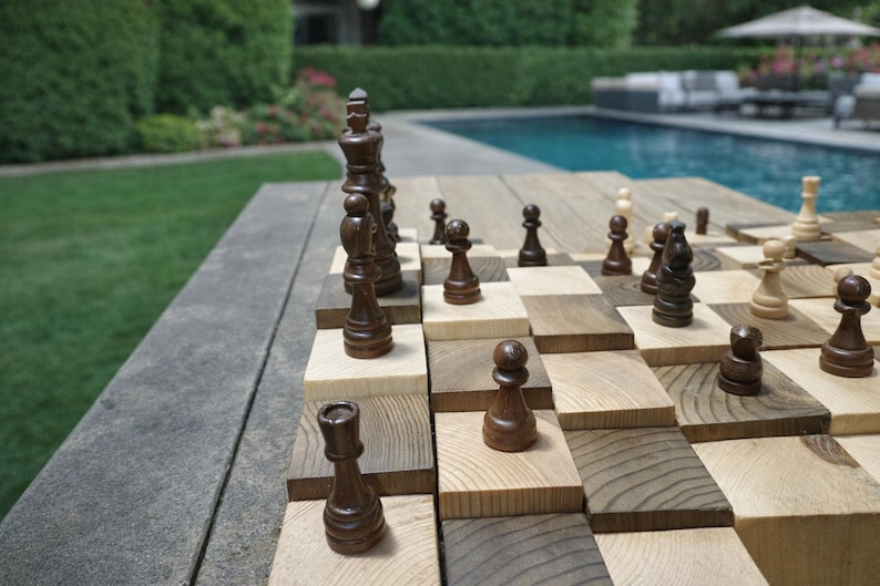 Puede incluir: Un juego de ajedrez de madera con piezas de madera marr&oacute;n oscuro y clara sobre un tablero &uacute;nico y elevado. El tablero est&aacute; instalado al aire libre cerca de una piscina y un c&eacute;sped verde. Las piezas de ajedrez est&aacute;n en las posiciones iniciales.