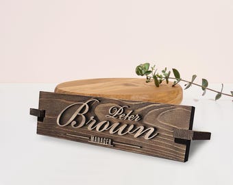 Op de afbeelding: Een houten naambord met de tekst "Peter Brown" en de titel "Manager" gegraveerd in lichtbruin. Het naambord ligt op een lichtbruin houten oppervlak.