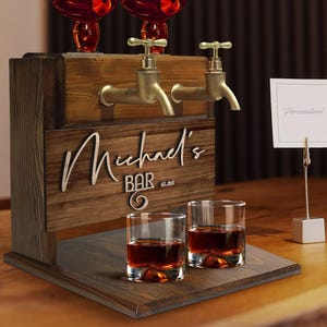 以下が含まれることがあります： 2つの金の注ぎ口と「Michael's Bar Est. 2020」と書かれたパーソナライズされたサインが付いた木製バーカート。カートの表面には、透明なガラス製のショットグラスが2つ置かれています。