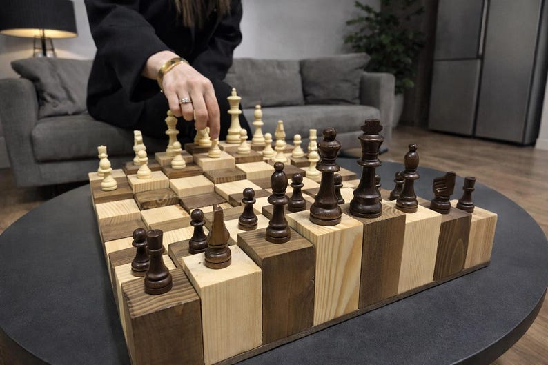 Puede incluir: Un juego de ajedrez de madera &uacute;nico con casillas claras y oscuras alternas. Las piezas del ajedrez son de color marr&oacute;n oscuro y beige claro. El tablero est&aacute; elevado, creando un efecto 3D. El juego est&aacute; sobre una mesa redonda y oscura.