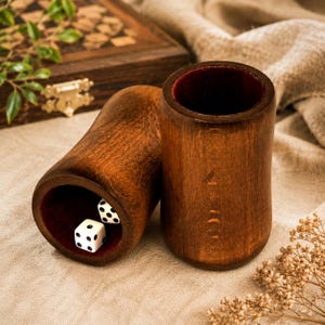 May include: Two cylindrical wooden dice cups with a dark brown finish and a deep red velvet lining. One cup stands upright, while the other lies on its side, revealing two white six-sided dice with black pips resting inside the velvet-lined interior.