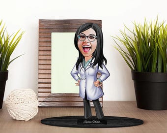 May include: A caricature figurine of a person wearing a white lab coat and glasses. The figurine is standing on a black base with the name "Sophia Miller" printed on it. The figurine is in front of a wooden picture frame.