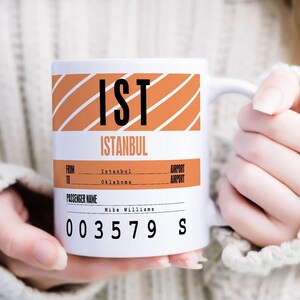 Op de afbeelding: Een witte keramische mok met oranje en witte strepen. De mok heeft de tekst "IST ISTANBUL" op de voorkant gedrukt. Onder de tekst is er een sectie voor "FROM TO" en "PASSENGER NAME". De mok heeft ook een sectie voor "AIRPORT AIRPORT". De mok heeft de nummers "003579" en de letter "S" op de bodem gedrukt.