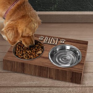 May include: A wooden pet food and water bowl stand with the name "Daisy" engraved on the front. The stand has two stainless steel bowls, one with dog food and the other with water. A golden retriever dog is eating from the food bowl.