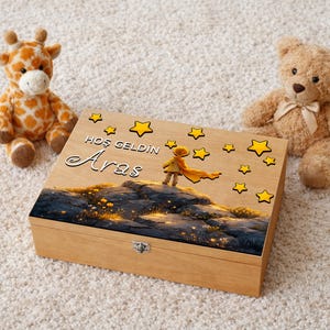 Pode incluir: Caixa de lembranças de madeira com tampa articulada, com uma ilustração pintada de uma criança com um lenço amarelo em uma paisagem rochosa. A caixa é adornada com estrelas amarelas e o texto "Hoş Geldin Aras". Pelúcias de girafa e urso de pelúcia em ambos os lados.